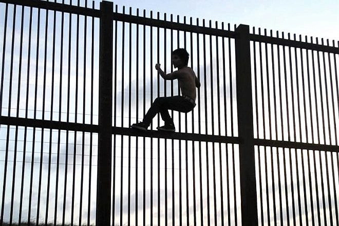 An immigrant attempting to cross the U.S.-Mexico border by illegally by climbing over the border fence in Brownsville, Texas in July 2009. Credit: Nofx221984 via Wikimedia Commons.