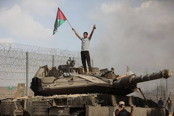 Palestinians wave flags and take photos next to a burning Israeli Defense Forces tank inside the border fence with Israel near the city of Khan Yunis in the southern Gaza Strip, Oct. 7, 2023. Photo by Yousef Mohammed/Flash90.